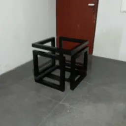 Czarna, metalowa konstrukcja przestrzenna przypominająca sześcian z przerwami, ustawiona na szarej podłodze w pomieszczeniu z brązowymi drzwiami w tle.