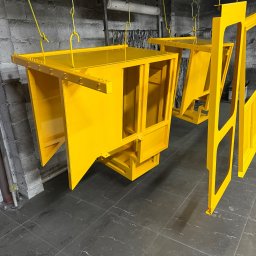 WELDING Łukasz Kułak - Żółte, metalowe elementy konstrukcyjne, prawdopodobnie kabiny maszyn, wiszą na hakach w warsztacie po malowaniu. Widoczne ślady użytkowania na podłodze.