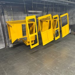 WELDING Łukasz Kułak - Żółte, metalowe elementy konstrukcyjne kabiny maszyny budowlanej, zawieszone na hakach w lakierni. Widoczne ślady użytkowania na podłodze.