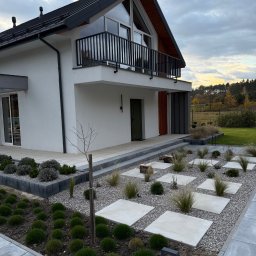 WELDING Łukasz Kułak - Nowoczesny dom z balkonem z czarną, metalową balustradą. Ogród z kamieniami i płytami chodnikowymi. Widok na elewację i zagospodarowanie terenu.