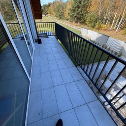 WELDING Łukasz Kułak - Nowoczesny balkon z czarną, metalową balustradą i szarymi płytkami, widok na zielony ogród i las w tle. Minimalistyczny design, jasne otoczenie, częściowo widoczna noga fotografa.