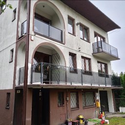 WELDING Łukasz Kułak - Dwupiętrowy dom z balkonami zabezpieczonymi balustradami szklanymi. Widoczne narzędzia sugerują prace wykończeniowe. Ujęcie z dołu, podkreślające architekturę budynku.