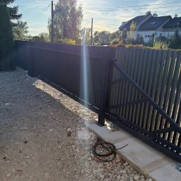 WELDING Łukasz Kułak - Nowoczesna, przesuwna brama wjazdowa z pionowymi panelami, zamontowana na betonowej podstawie. Widoczne okablowanie sugeruje automatykę. Tło: dom jednorodzinny.