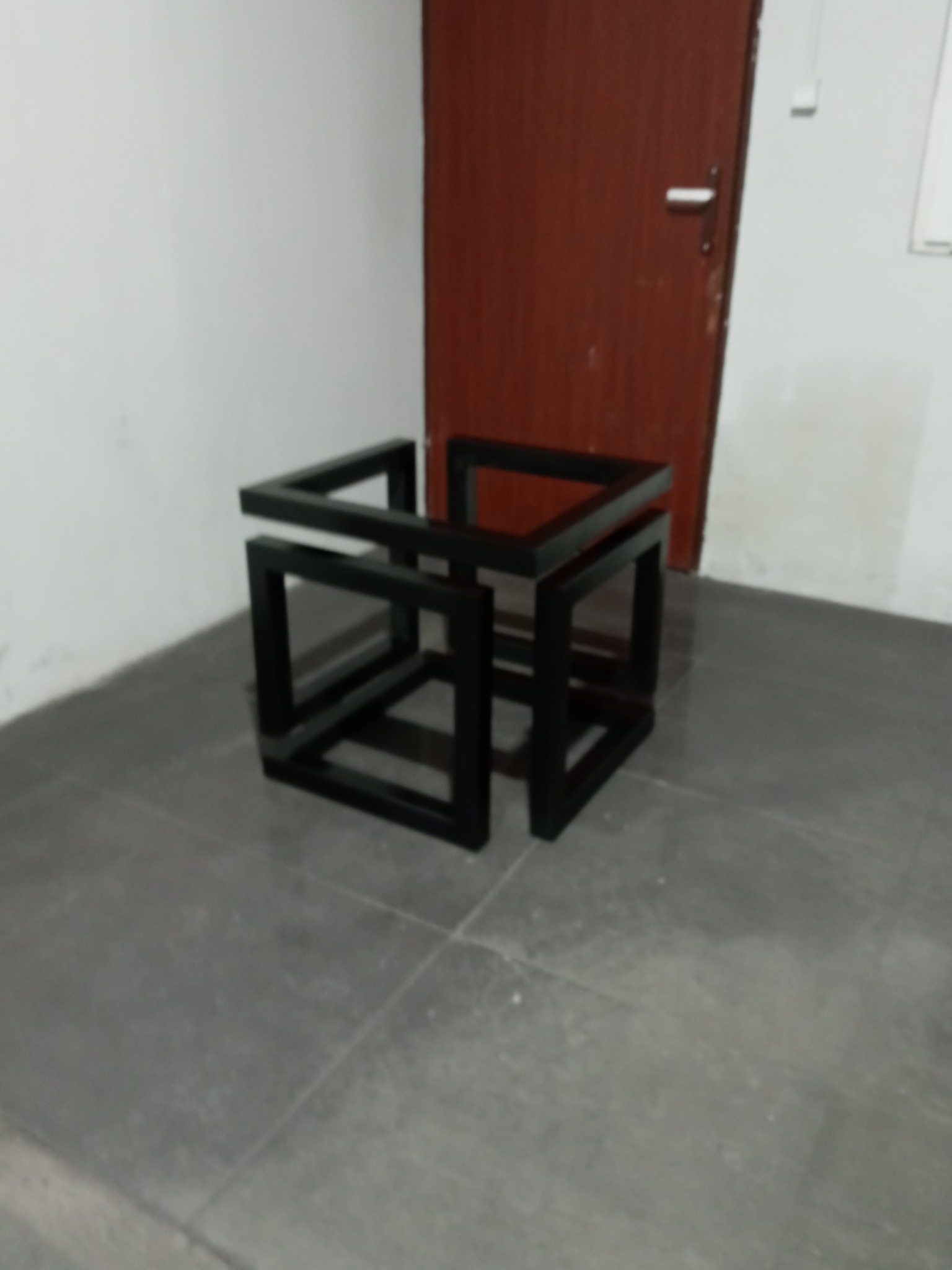 Czarna, metalowa konstrukcja przestrzenna przypominająca sześcian z przerwami, ustawiona na szarej podłodze w pomieszczeniu z brązowymi drzwiami w tle.