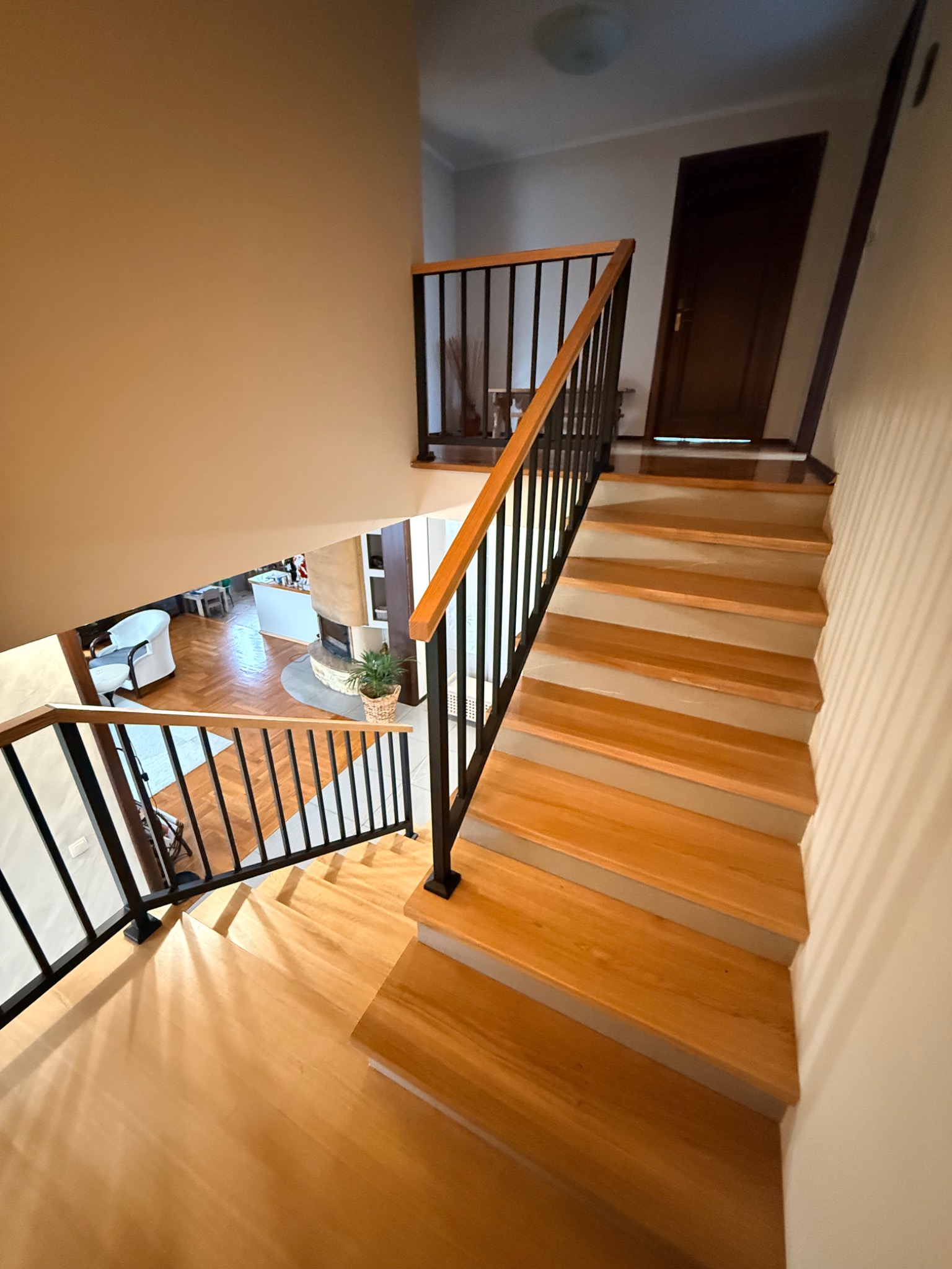 Drewniane schody z czarną balustradą, widok z góry. Jasne drewno stopni kontrastuje z ciemnymi elementami balustrady. W tle widoczny salon z kominkiem.
