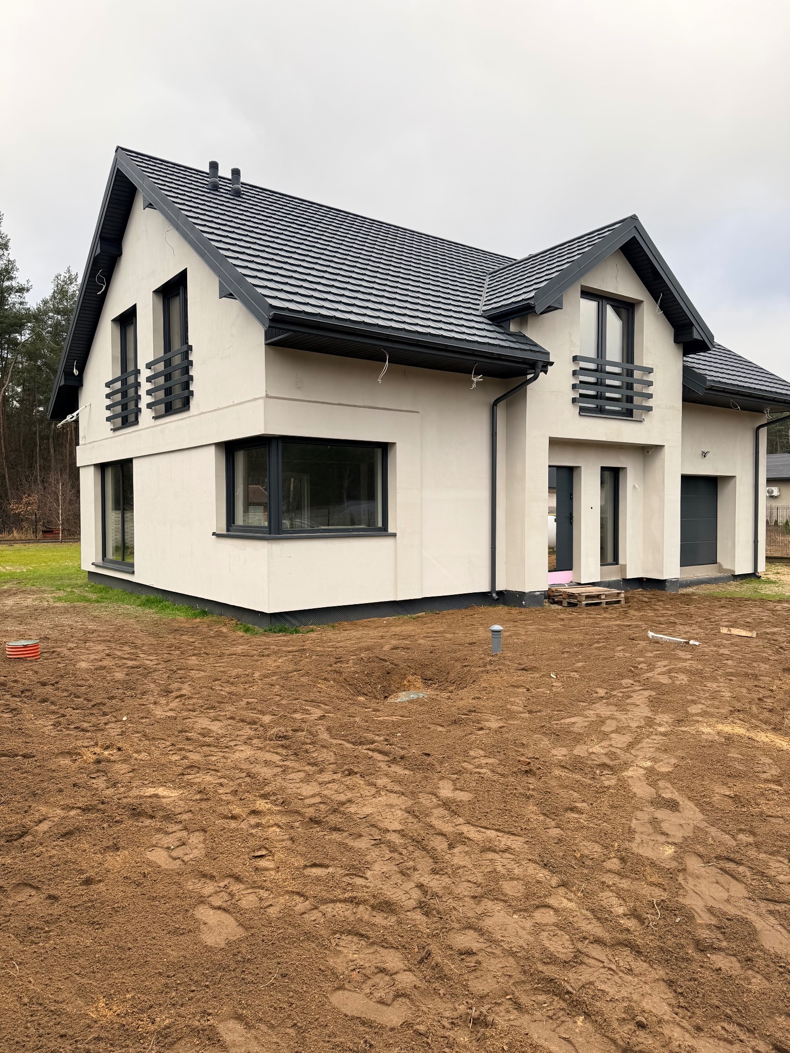 Nowoczesny dom jednorodzinny z szarą dachówką i elementami wykończeniowymi w kolorze antracytowym, widok z zewnątrz na przygotowanym terenie pod trawnik.