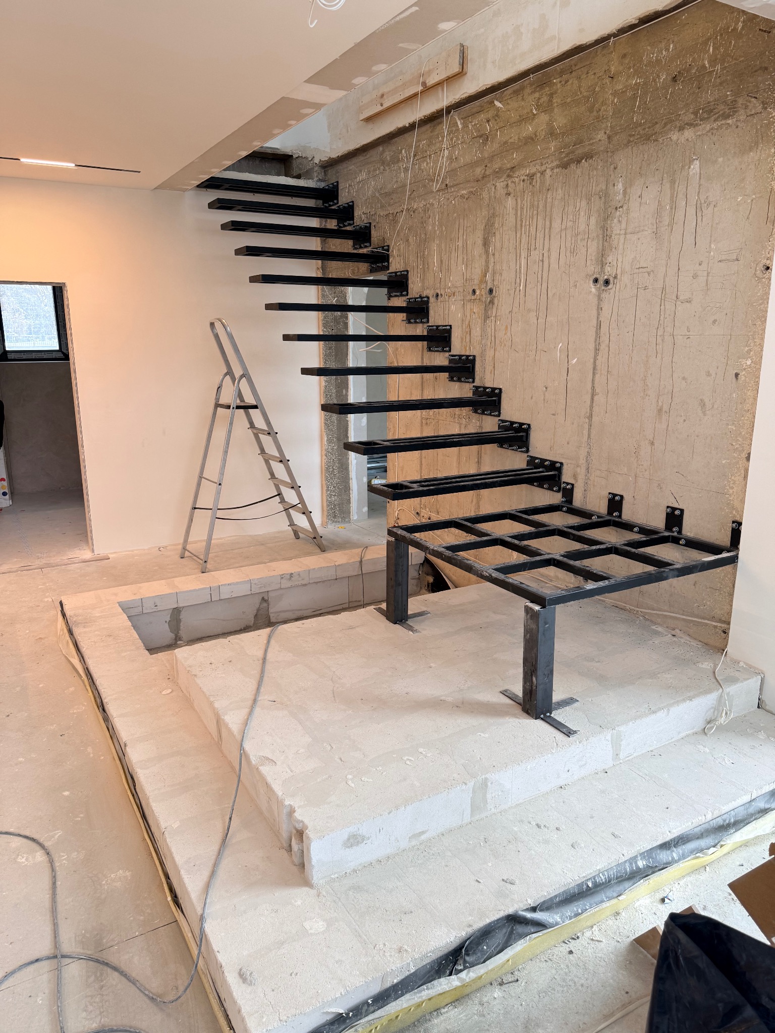 Surowe, metalowe schody modułowe w trakcie montażu w surowym wnętrzu. Widoczna drabina i betonowe podłoże. Elementy spawane, mocowane do ściany.
