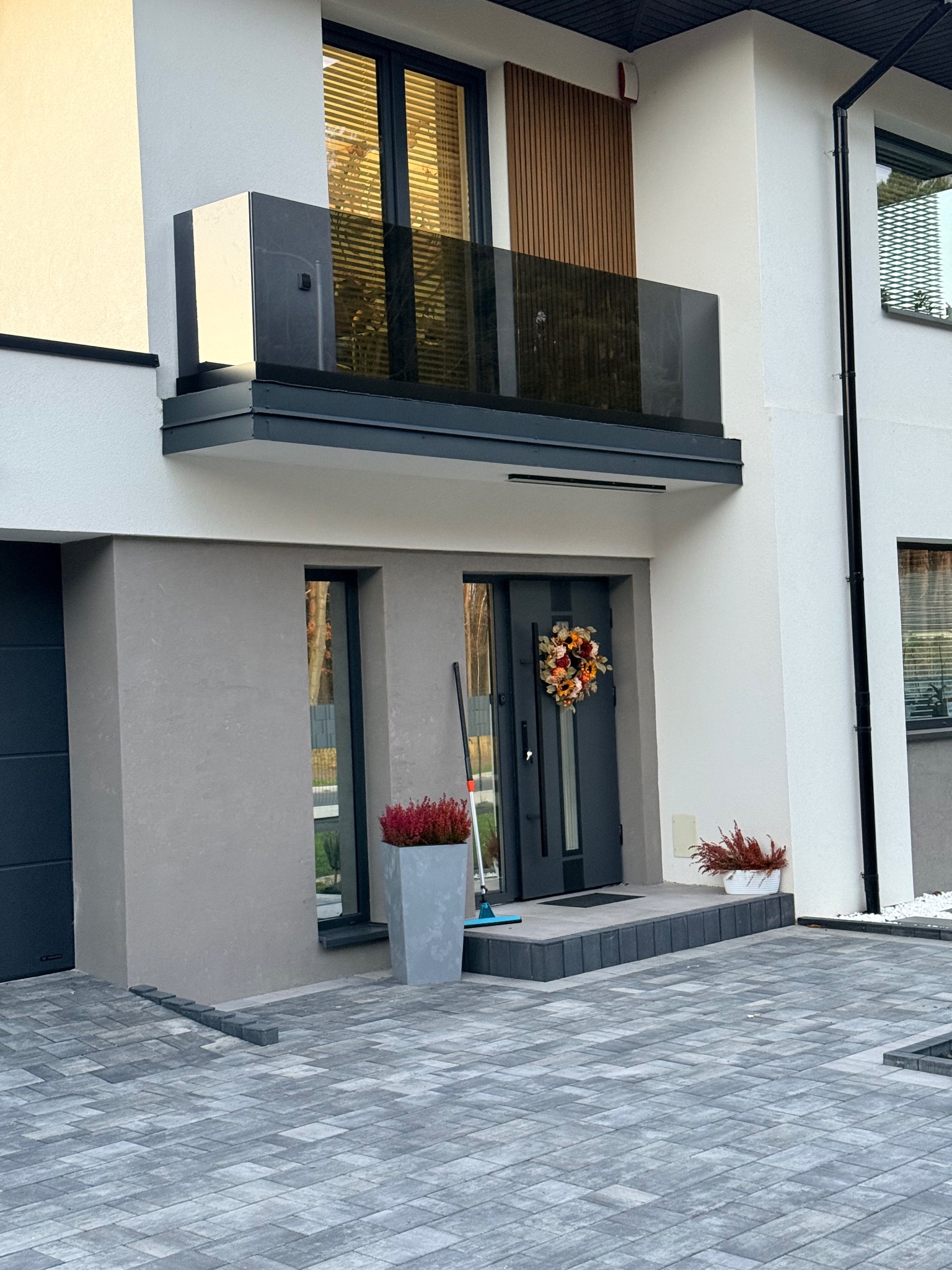 Nowoczesny dom z szarą elewacją, balkonem z ciemną szklaną balustradą i wejściem ozdobionym jesiennym wieńcem. Kostka brukowa na podjeździe.