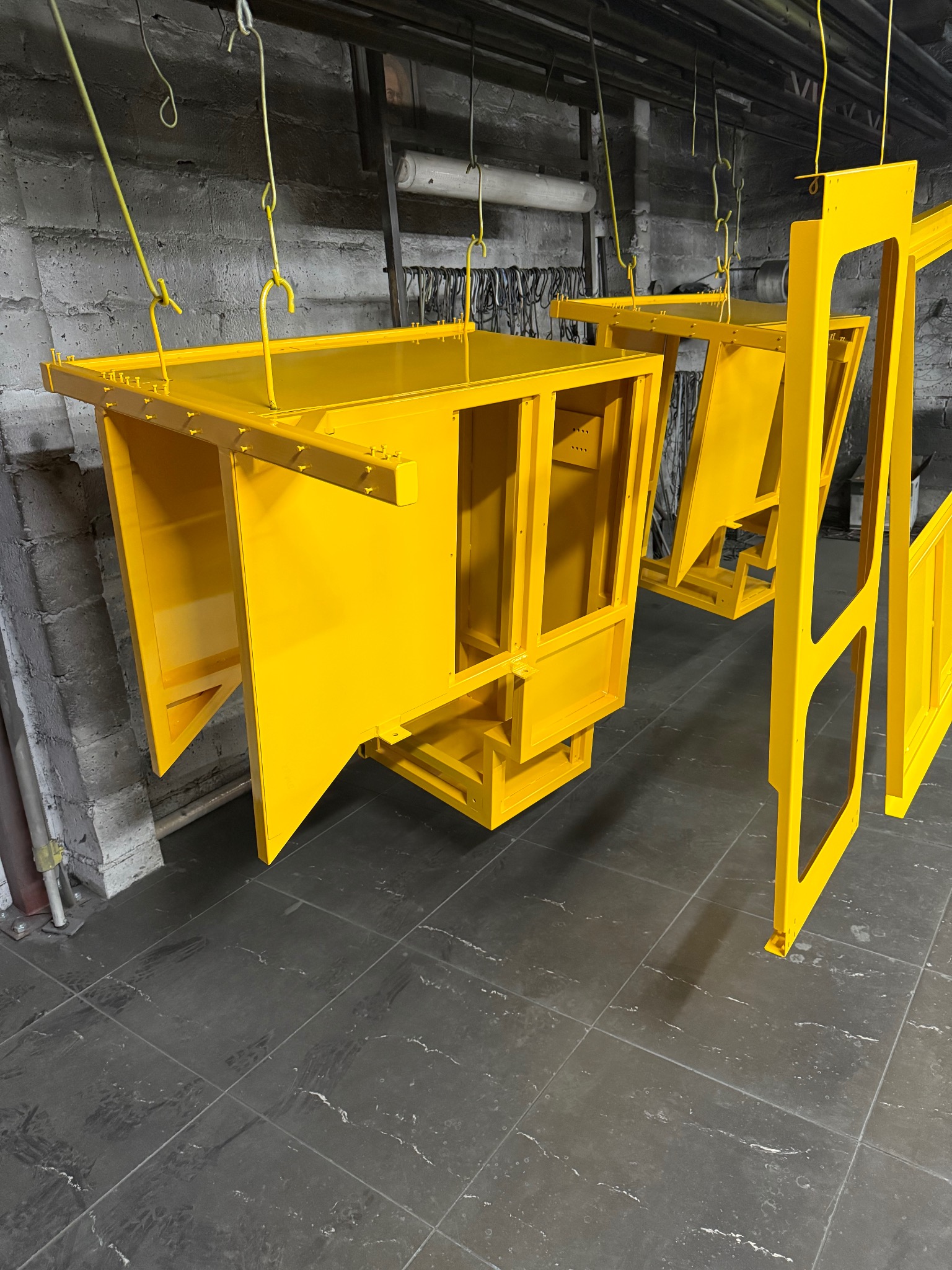 Żółte, metalowe elementy konstrukcyjne, prawdopodobnie kabiny maszyn, wiszą na hakach w warsztacie po malowaniu. Widoczne ślady użytkowania na podłodze.
