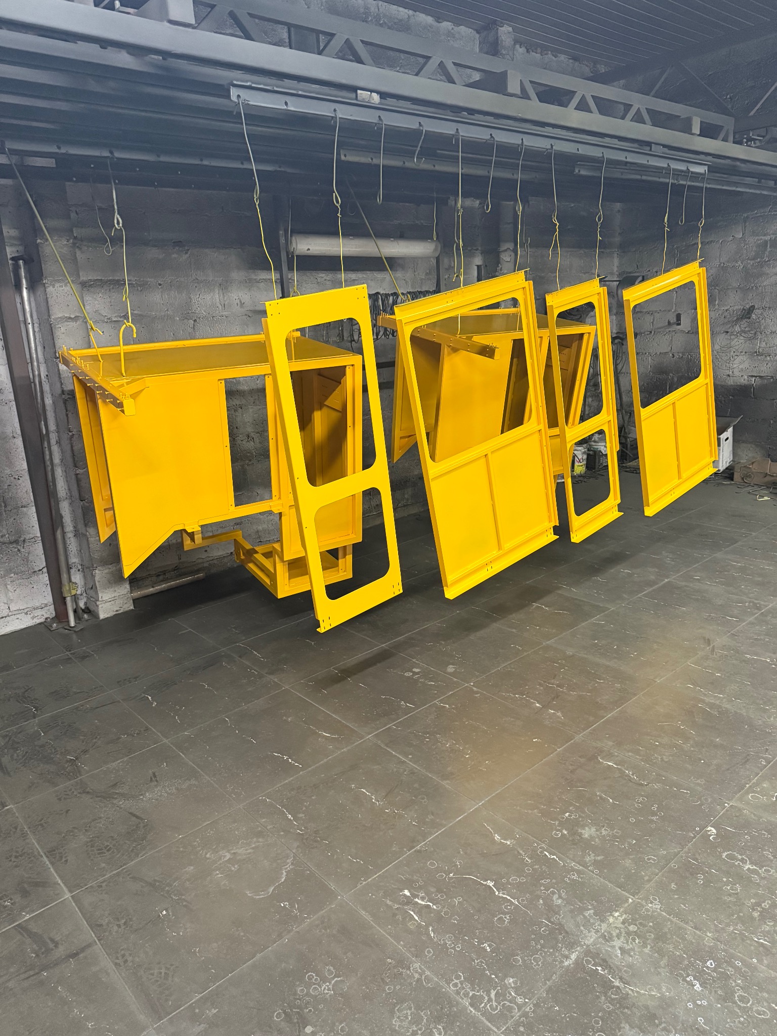 Żółte, metalowe elementy konstrukcyjne kabiny maszyny budowlanej, zawieszone na hakach w lakierni. Widoczne ślady użytkowania na podłodze.