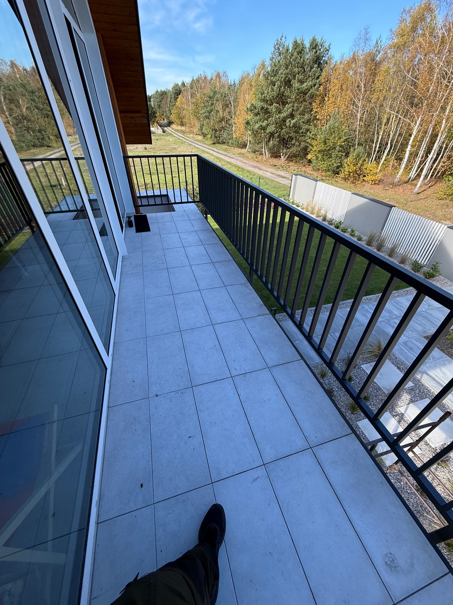 Nowoczesny balkon z czarną, metalową balustradą i szarymi płytkami, widok na zielony ogród i las w tle. Minimalistyczny design, jasne otoczenie, częściowo widoczna noga fotografa.
