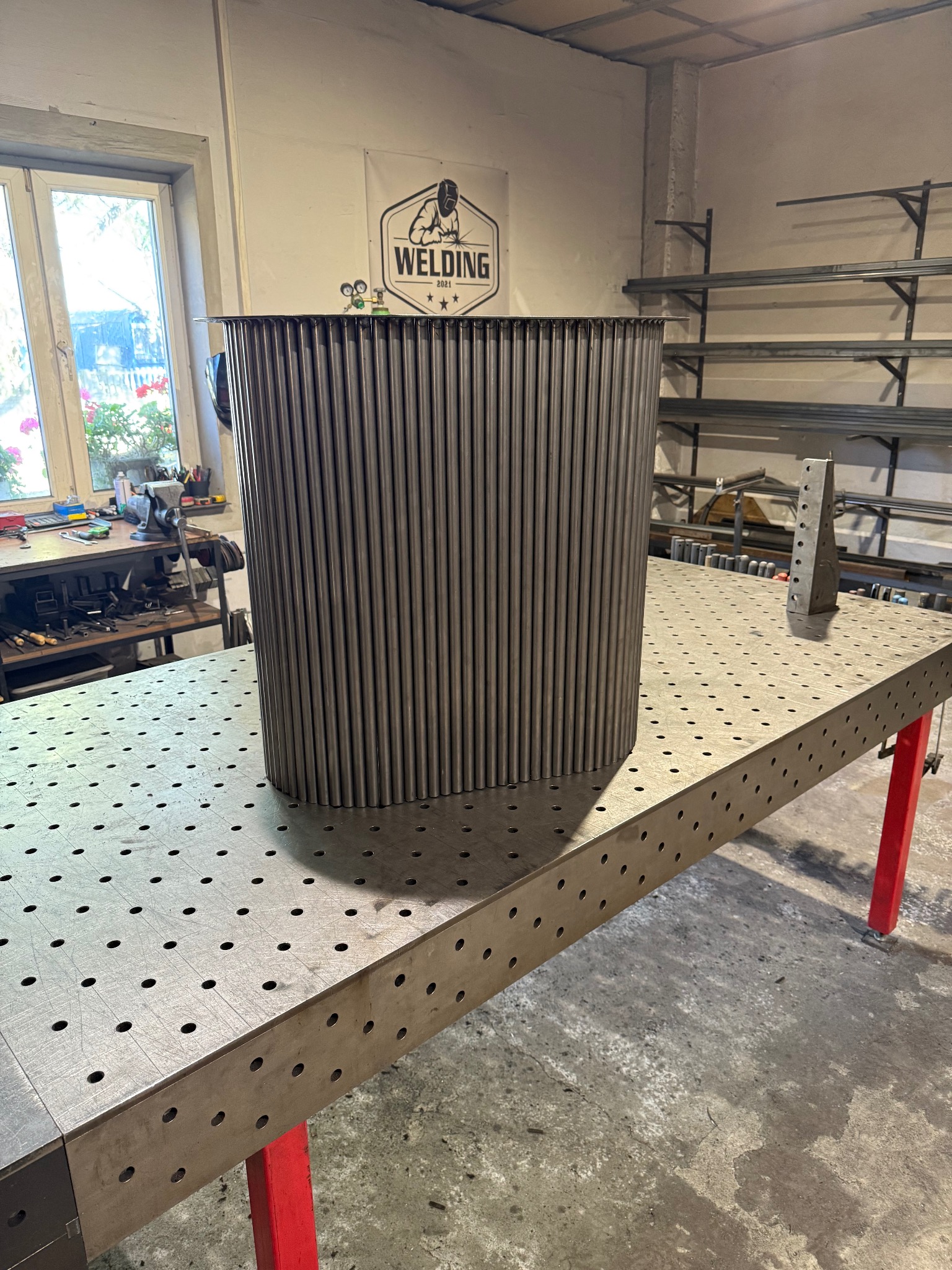 Metalowa konstrukcja z rurek na perforowanym stole spawalniczym w warsztacie, widoczne narzędzia i logo firmy spawalniczej w tle.