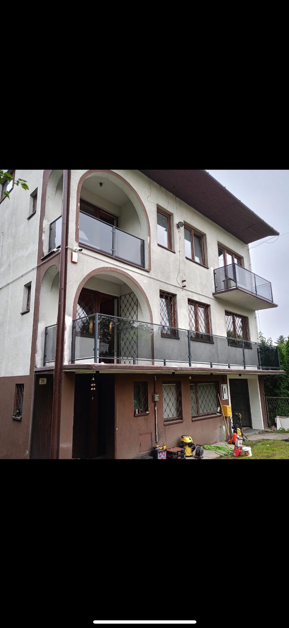 Dwupiętrowy dom z balkonami zabezpieczonymi balustradami szklanymi. Widoczne narzędzia sugerują prace wykończeniowe. Ujęcie z dołu, podkreślające architekturę budynku.