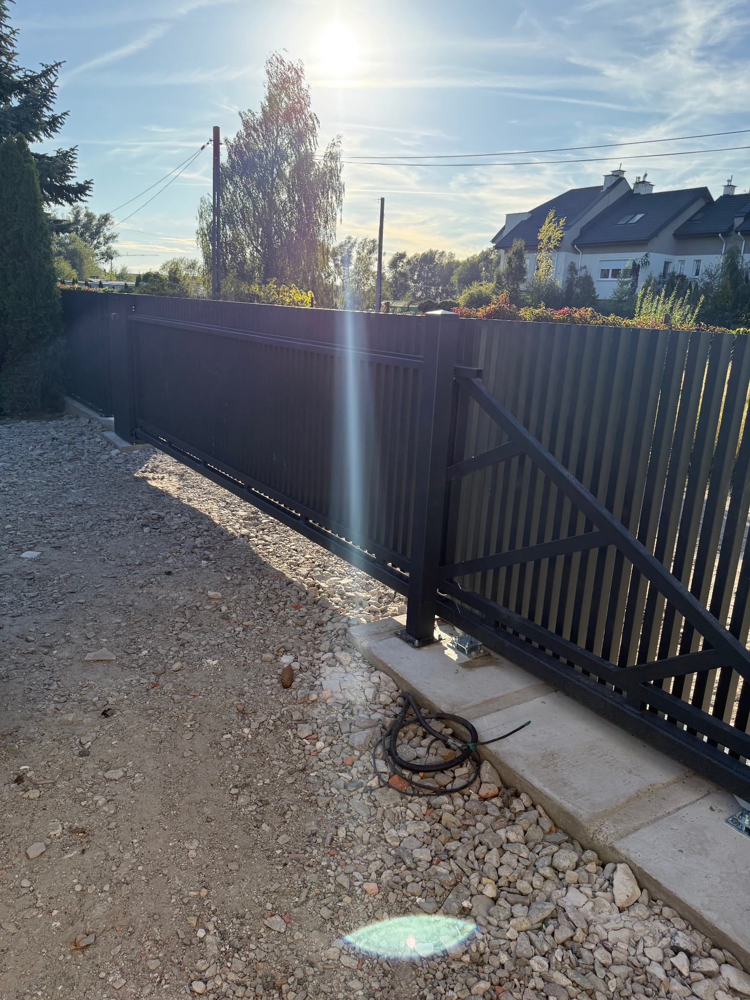 Nowoczesna, przesuwna brama wjazdowa z pionowymi panelami, zamontowana na betonowej podstawie. Widoczne okablowanie sugeruje automatykę. Tło: dom jednorodzinny.