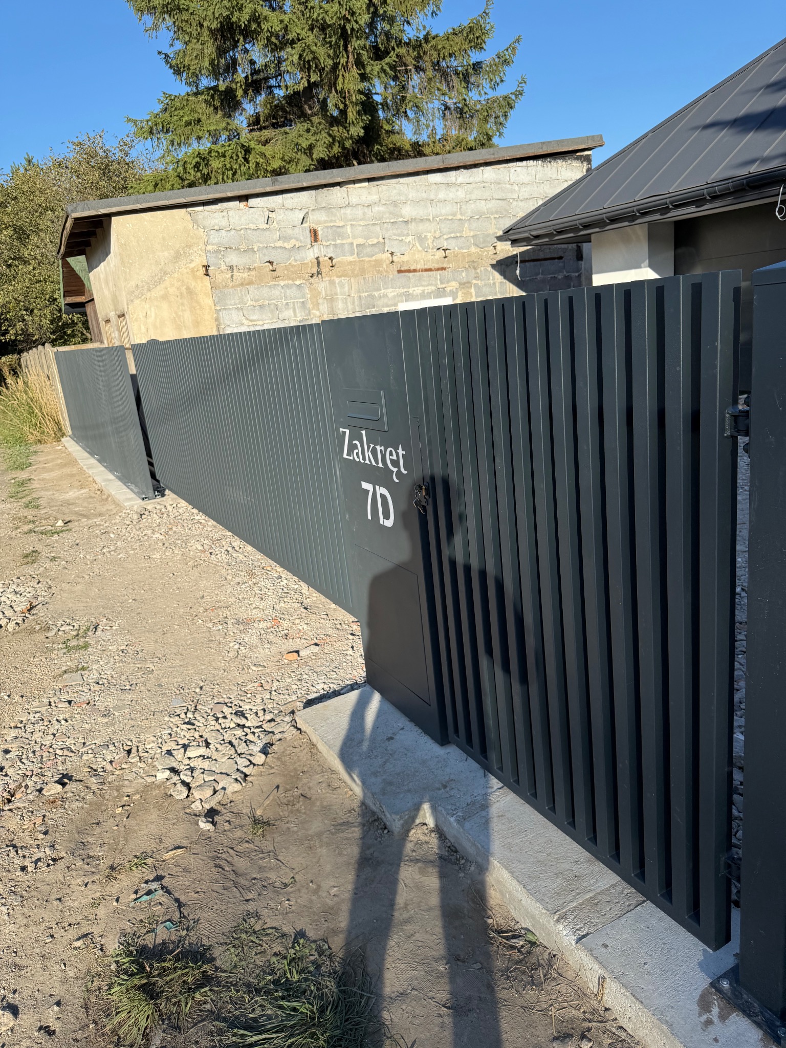 Nowoczesna, grafitowa brama przesuwna z adresem 'Zakręt 7D', z pionowymi panelami, na tle budynku i żwirowego podłoża. Widoczny cień osoby.
