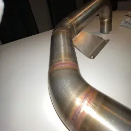 Metalowa rura wydechowa z widocznymi spawami i uchwytem mocującym, leżąca na białym stole. Widoczne ślady obróbki termicznej na spoinach.