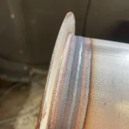 Szczegółowe zbliżenie na spawany element metalowy, widoczne ślady po spawaniu z charakterystycznymi tęczowymi przebarwieniami termicznymi.