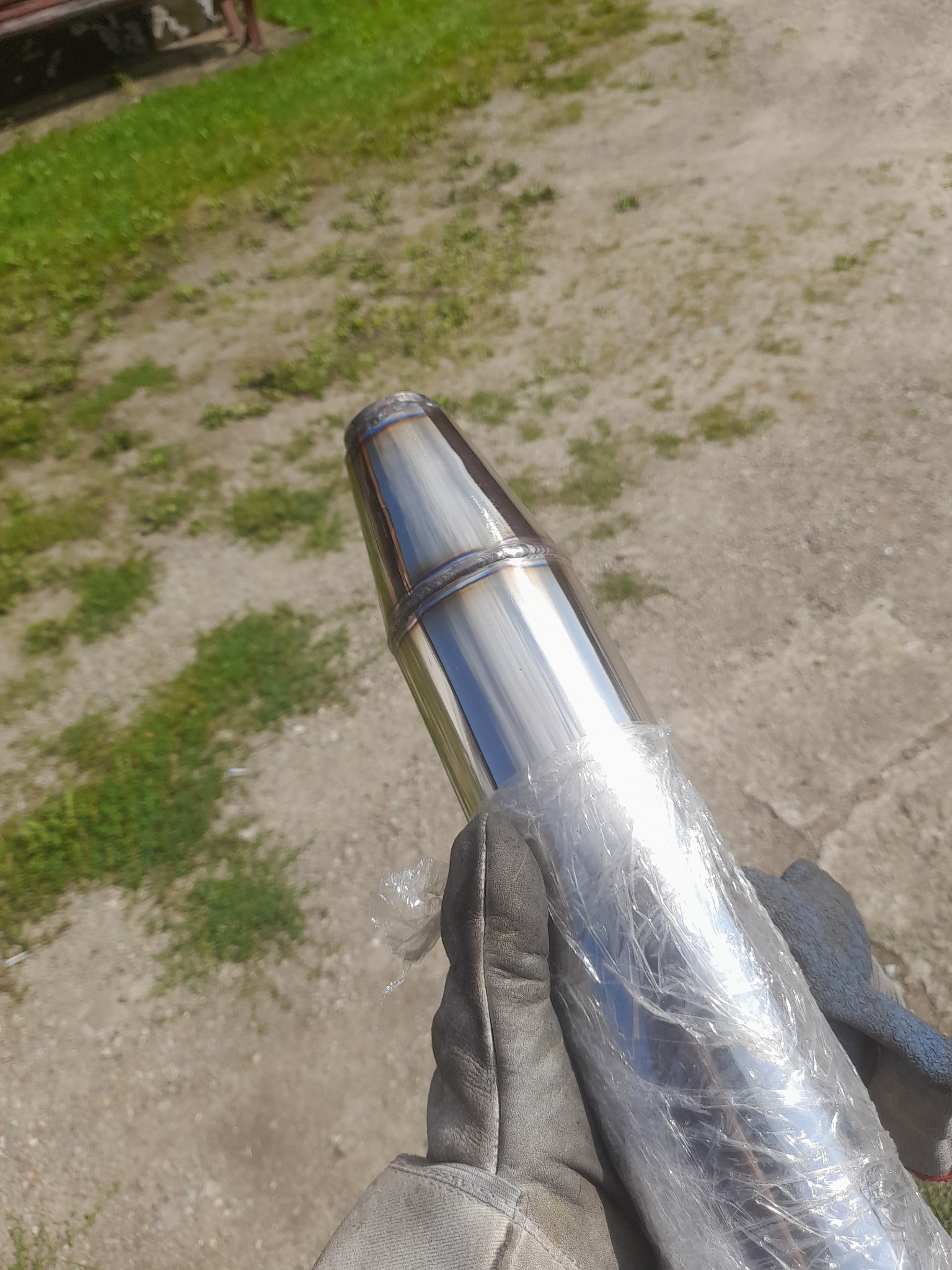 Błyszczący, spawany element metalowy, prawdopodobnie rura wydechowa, owinięty folią ochronną i trzymany w rękawicy spawalniczej na tle trawy i ziemi.