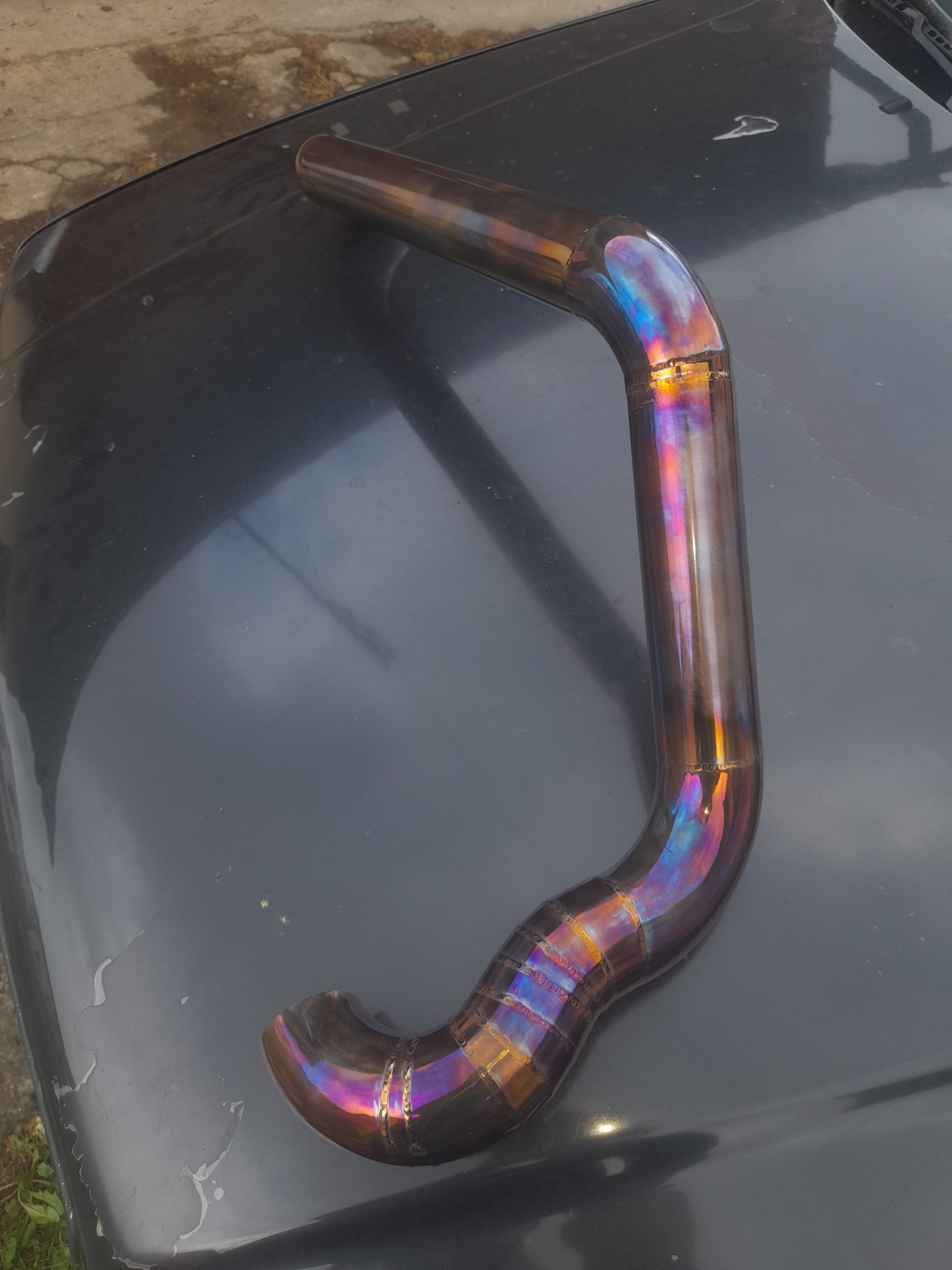 Metalowa rura o zakrzywionym kształcie, z widocznymi śladami spawania w postaci regularnych zgrubień, prezentująca tęczowe zabarwienie na powierzchni, leżąca na czarnej, lekko zabrudzonej powierzchni.