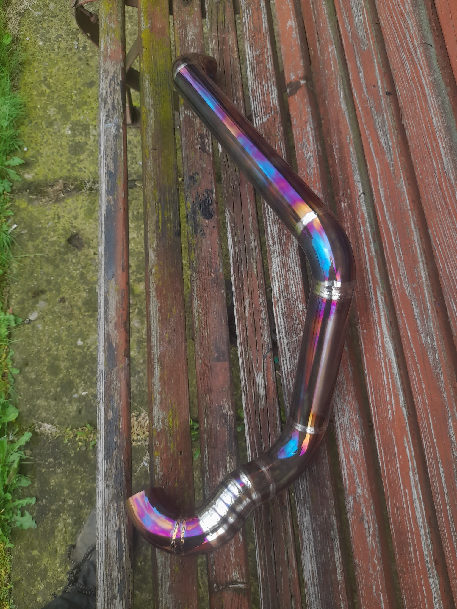 Wygięta, metalowa rura z widocznymi śladami spawania, opalizująca kolorami tęczy, leżąca na starej, drewnianej ławce.