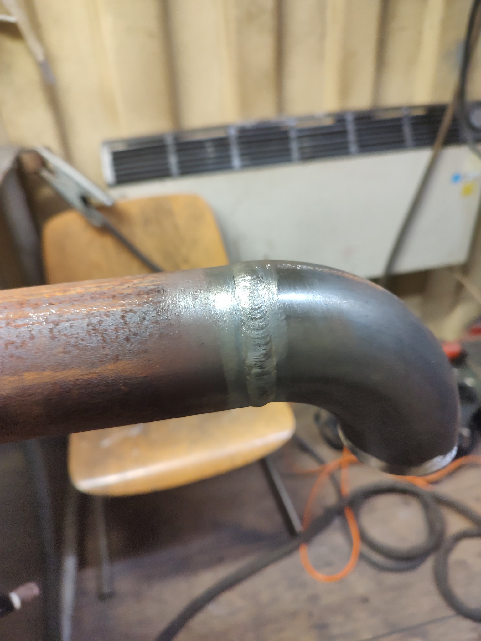 Zespawany fragment rury stalowej z widocznym łączeniem spawalniczym, część rury pokryta rdzą, w tle widoczne narzędzia i elementy wyposażenia warsztatu.