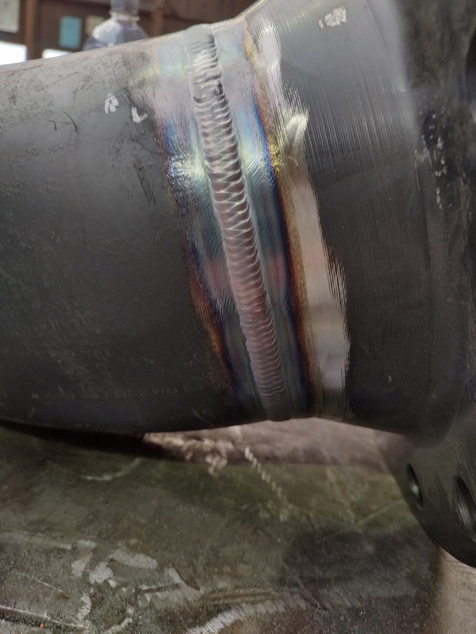 Spoina na rurze z widocznymi kolorami hartowania, wykonana na ciemnoszarym metalu.