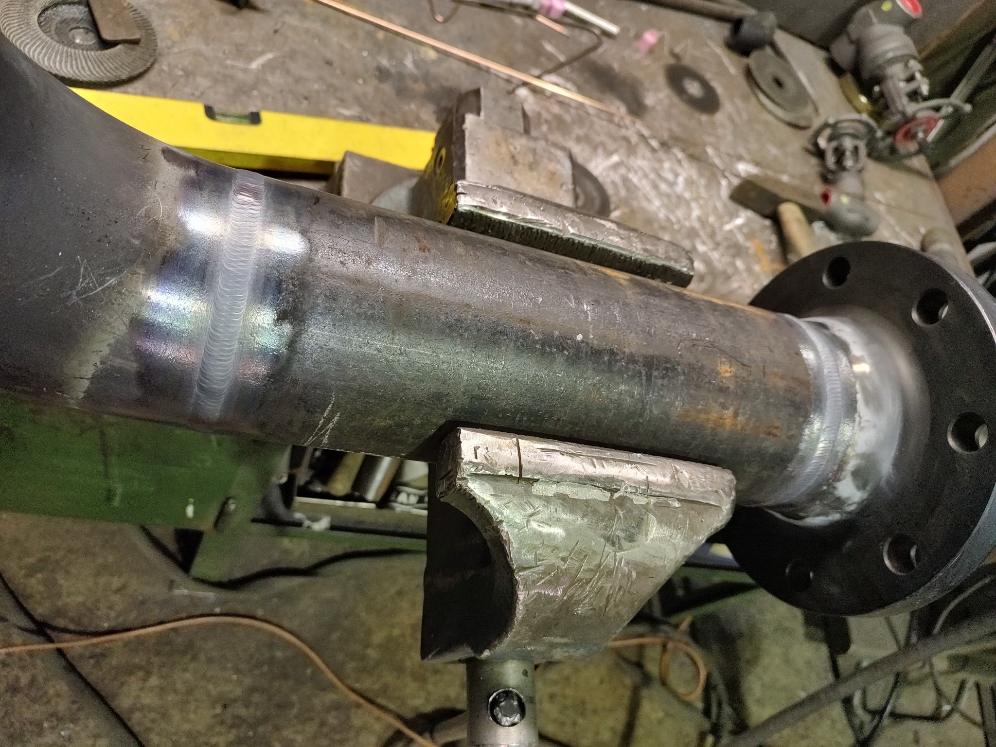 Spoina na rurze z kołnierzem, zamocowanej w metalowym uchwycie na warsztatowym stole, widoczne ślady obróbki i narzędzia w tle.