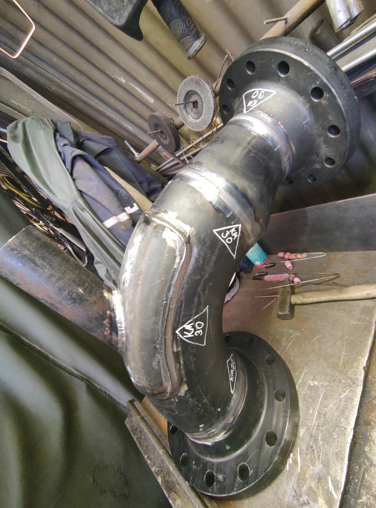 Zespawany element rurociągu z kołnierzami, z widocznymi śladami spawania i oznaczeniami 'KM 30', leżący na metalowym stole warsztatowym, obok narzędzi spawalniczych.