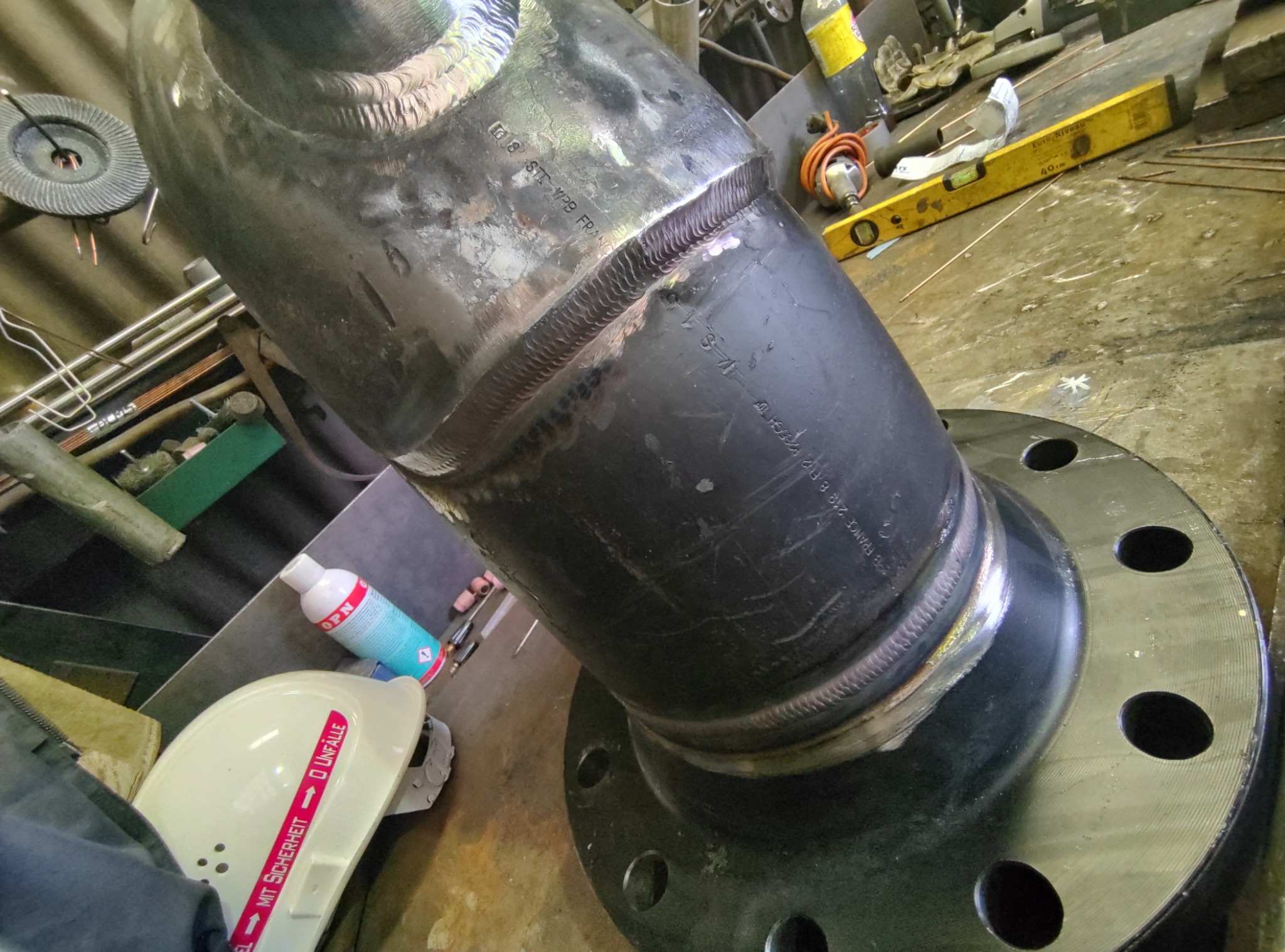 Spoina na rurze stalowej z kołnierzem, widoczne ślady po obróbce, obok kask ochronny z naklejką ostrzegawczą i butelka sprayu.