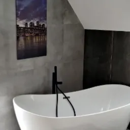 Wolnostojąca wanna z czarną armaturą na tle szarych płytek i obrazu z panoramą miasta w nowoczesnej łazience na poddaszu.