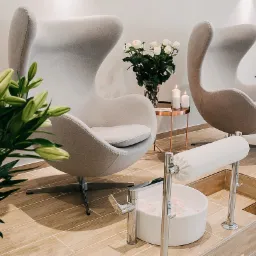 Dwa fotele typu Egg w jasnoszarym odcieniu, przygotowane do zabiegu na stopy, z misami do moczenia stóp z płatkami róż, bukietem białych róż i lilii w tle, oraz zapalonymi świecami na miedzianym...