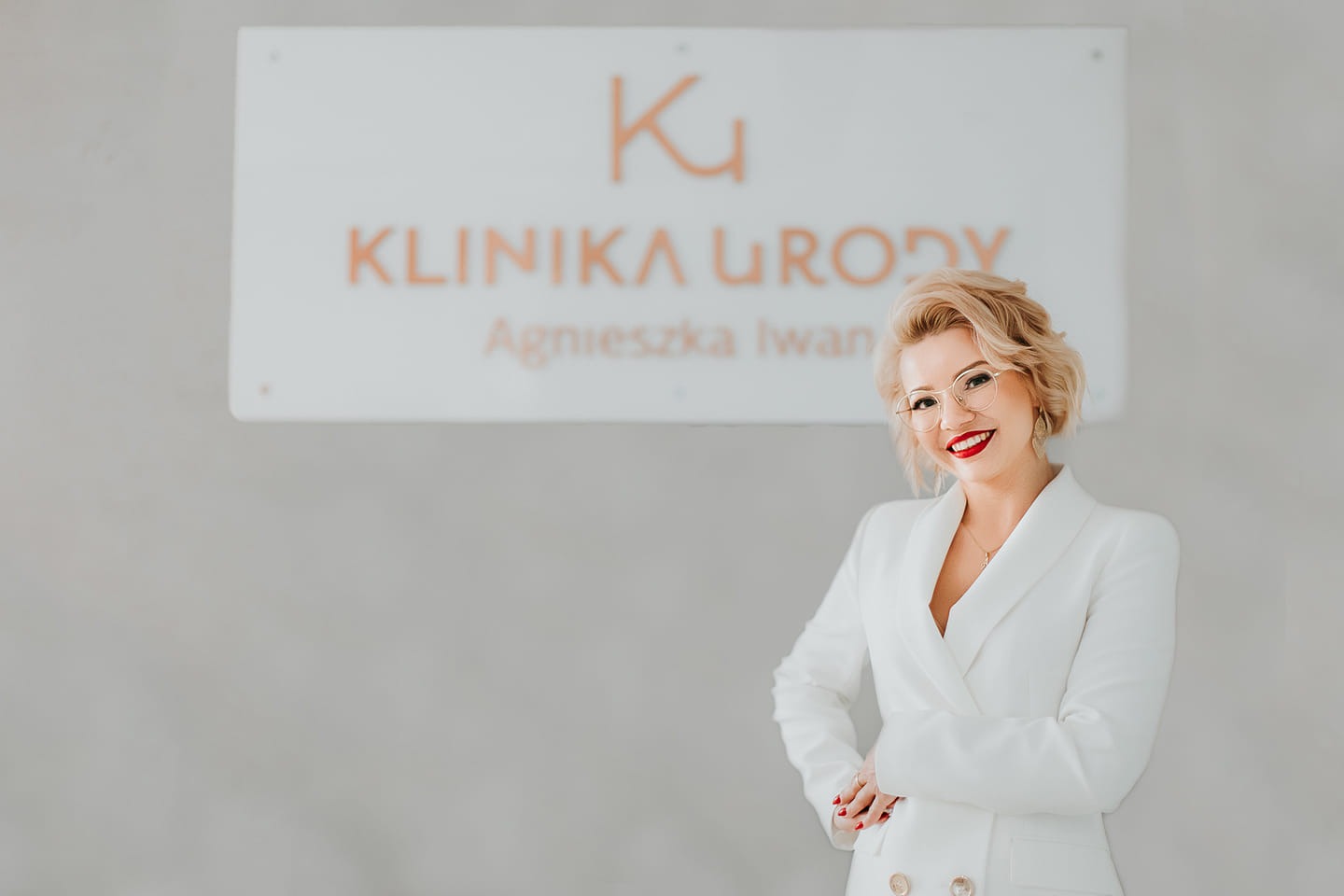 Portret uśmiechniętej właścicielki w białym żakiecie na tle szyldu Kliniki Urody Agnieszka Iwan w Ostrowcu Świętokrzyskim.