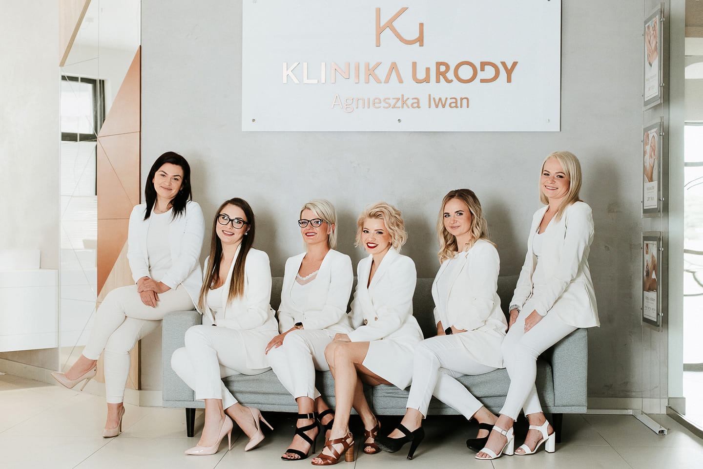 Zespół 'Kliniki Urody Agnieszka Iwan' w białych garniturach siedzi na szarej sofie na tle ściany z logo kliniki i dekoracyjnym lustrem.
