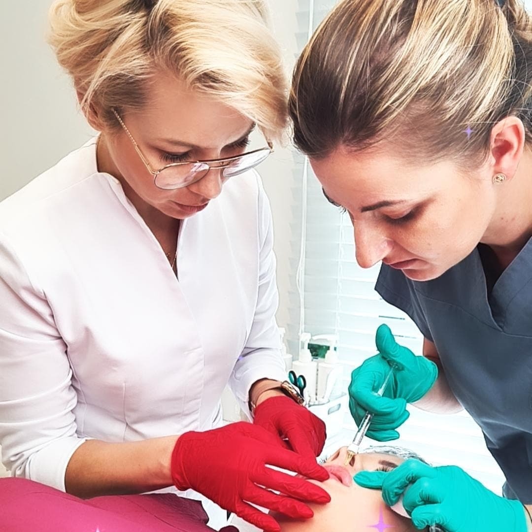 Dwie specjalistki w rękawicach (czerwonych i zielonych) wykonują zabieg iniekcji w usta pacjentki leżącej na różowym łóżku zabiegowym, w tle białe ściany gabinetu.