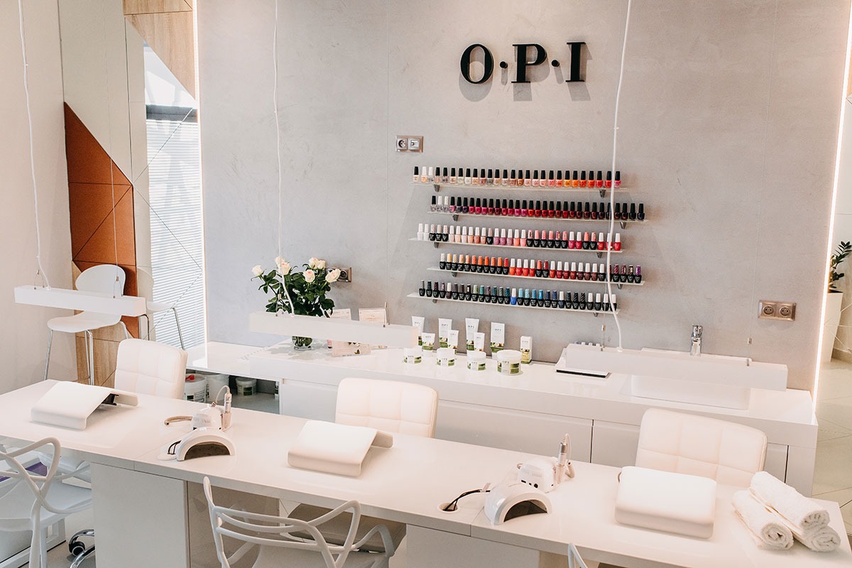 Jasne, przestronne wnętrze gabinetu z białymi stanowiskami do manicure, na ścianie półki z lakierami OPI, wazon z białymi różami i miękkie ręczniki.
