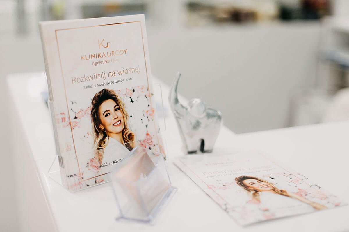 Białe biurko z ulotkami Kliniki Urody Agnieszki Iwan, z hasłem 'Rozkwitnij na wiosnę!' i ilustracją uśmiechniętej kobiety w kwiatach, obok srebrna figurka słonia.