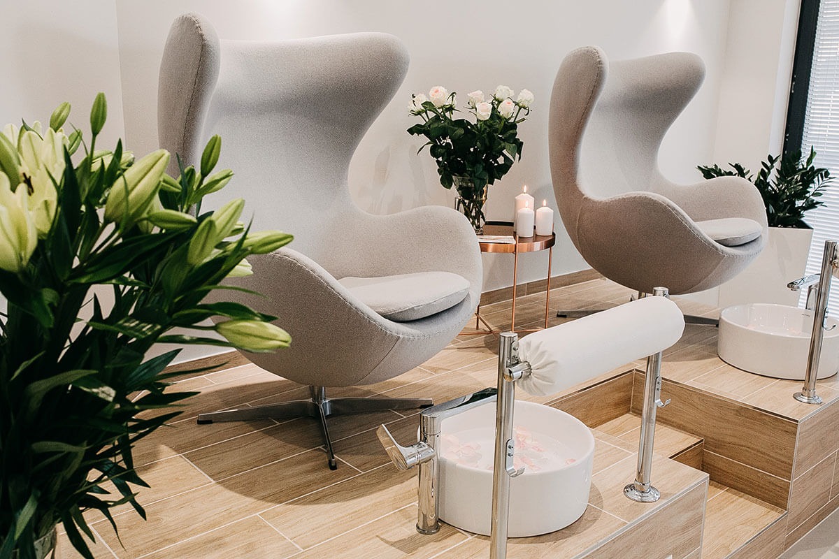 Dwa fotele typu Egg w jasnoszarym odcieniu, przygotowane do zabiegu na stopy, z misami do moczenia stóp z płatkami róż, bukietem białych róż i lilii w tle, oraz zapalonymi świecami na miedzianym...