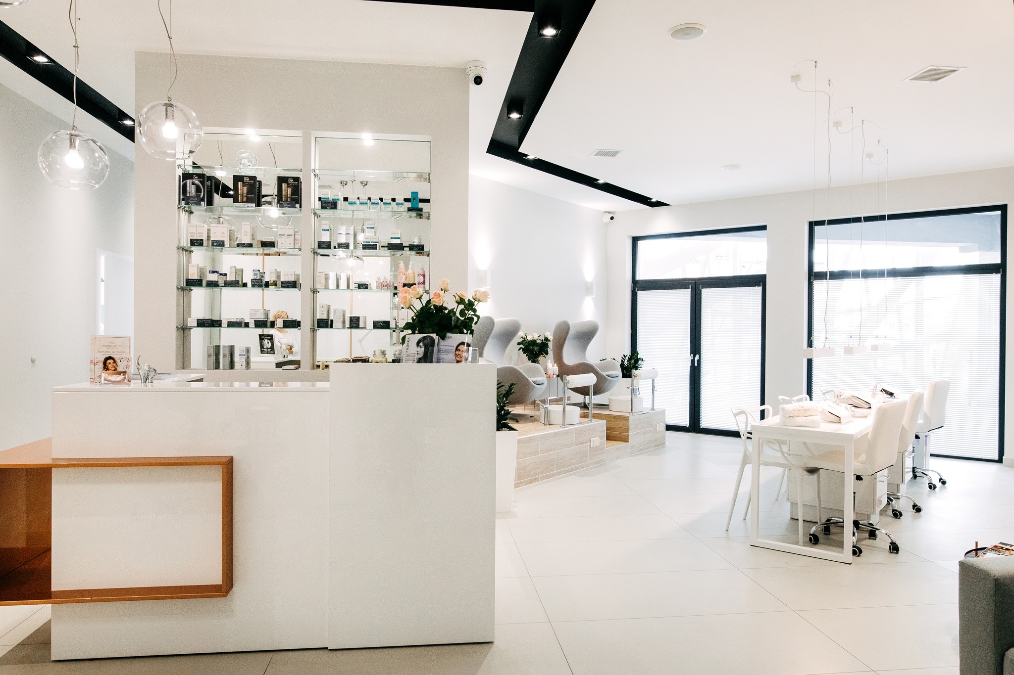 Jasne, przestronne wnętrze z recepcją z ekspozycją kosmetyków, wygodnymi fotelami dla klientów i stanowiskami do stylizacji paznokci, oświetlone wiszącymi lampami.