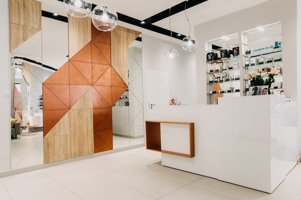 Nowoczesne wnętrze recepcji z geometryczną ścianą z luster i paneli drewnianych w odcieniach brązu, biała lada recepcyjna z drewnianymi akcentami, szklane półki z kosmetykami i bukiet róż w tle...
