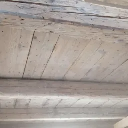 Drewniany sufit z belkami, widoczne ślady po gwoździach i naturalne przebarwienia drewna, w tle fragmenty konstrukcji budynku.