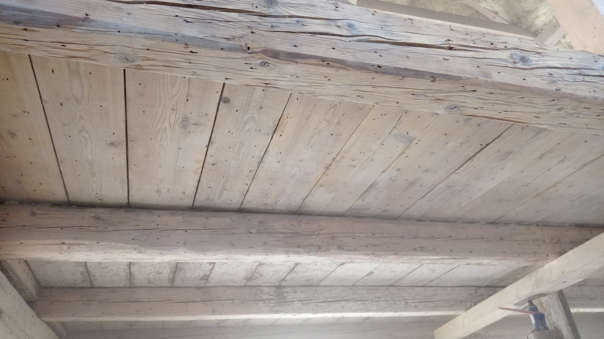 Drewniany sufit z belkami, widoczne ślady po gwoździach i naturalne przebarwienia drewna, w tle fragmenty konstrukcji budynku.