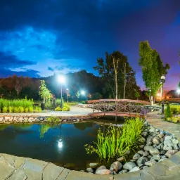 Panoramiczny widok na oświetlony park nocą z oczkiem wodnym obłożonym kamieniami, drewnianym mostkiem i ścieżką z kamiennych płyt.