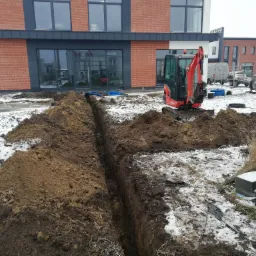 Wąski wykop w ziemi z widocznymi kablami, obok czerwona mini-koparka Kubota i budynek w tle, teren częściowo pokryty śniegiem.