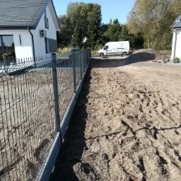 Nowo postawione, ocynkowane, grafitowe ogrodzenie z paneli na betonowej podmurówce, widok z zewnątrz posesji, z widocznymi śladami opon pojazdów budowlanych na ziemi i zaparkowanym busem firmy w tle.