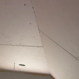 Skośny sufit poddasza z zamontowanymi płytami gipsowo-kartonowymi, widoczne wkręty, szpachlowane łączenia, taśma malarska przy ścianie, okno w tle.