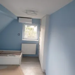 Pok&oacute;j na poddaszu po malowaniu na jasny niebieski kolor, z białymi panelami podłogowymi, grzejnikiem, klimatyzatorem i lampą sufitową w kształcie pierścieni, częściowo zasłonięty kartonem na podłodze.