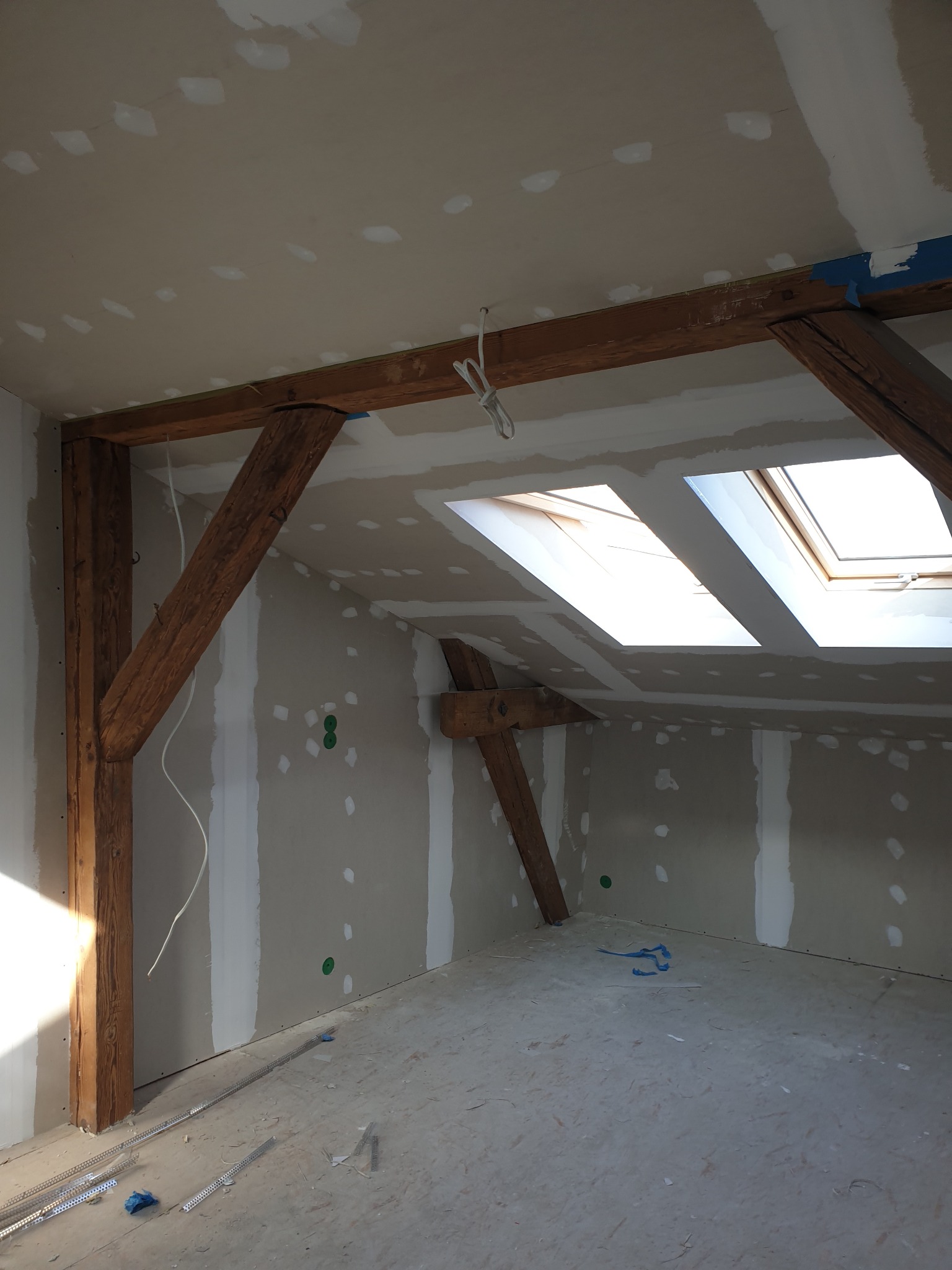 Poddasze w trakcie wykańczania: płyty karton-gips z widocznymi łączeniami szpachlowanymi, drewniane belki konstrukcyjne, okna dachowe wpuszczające światło, elementy montażowe na podłodze.