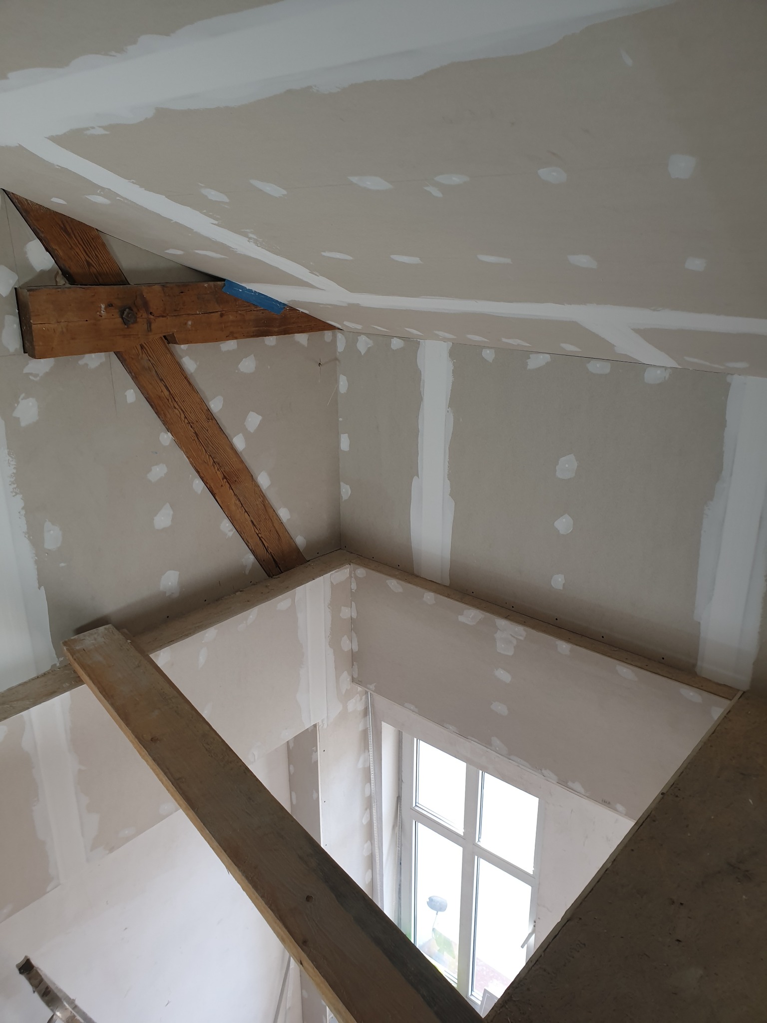 Poddasze z widocznymi drewnianymi belkami i zamontowanymi płytami karton-gips, szpachlowanymi łączeniami, widok na okno z dołu.