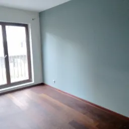 Pomalowane na jasnozielono ściany w pustym pokoju z drewnianą podłogą i dużym oknem balkonowym, widoczny plastikowy worek na podłodze.