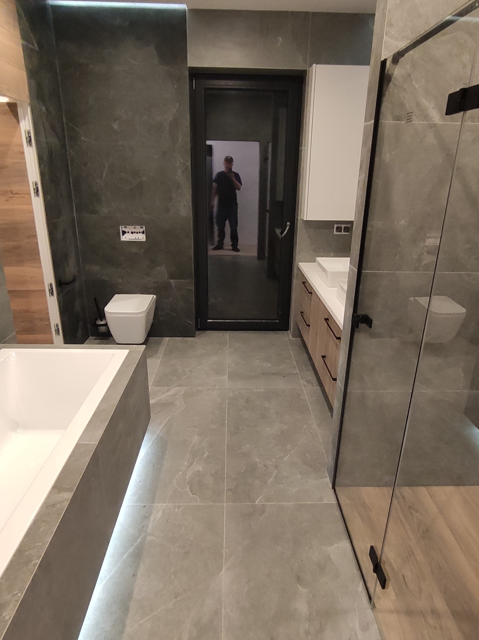 Nowoczesna łazienka z szarymi płytkami imitującymi marmur, podświetlaną wanną, kabiną prysznicową ze szkła, wiszącą szafką z umywalką i toaletą. W lustrze odbija się osoba robiąca zdjęcie.